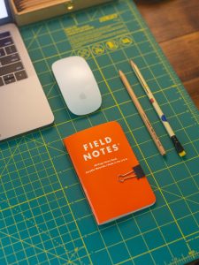 A desk with pencils and small field notes notebook. Computer and mouse. A workspace.