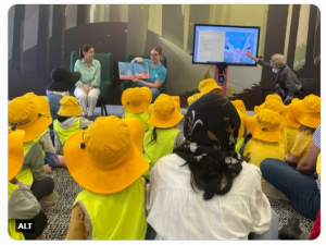 Two QUT students read a story to young children wearing yellow hats and vests.